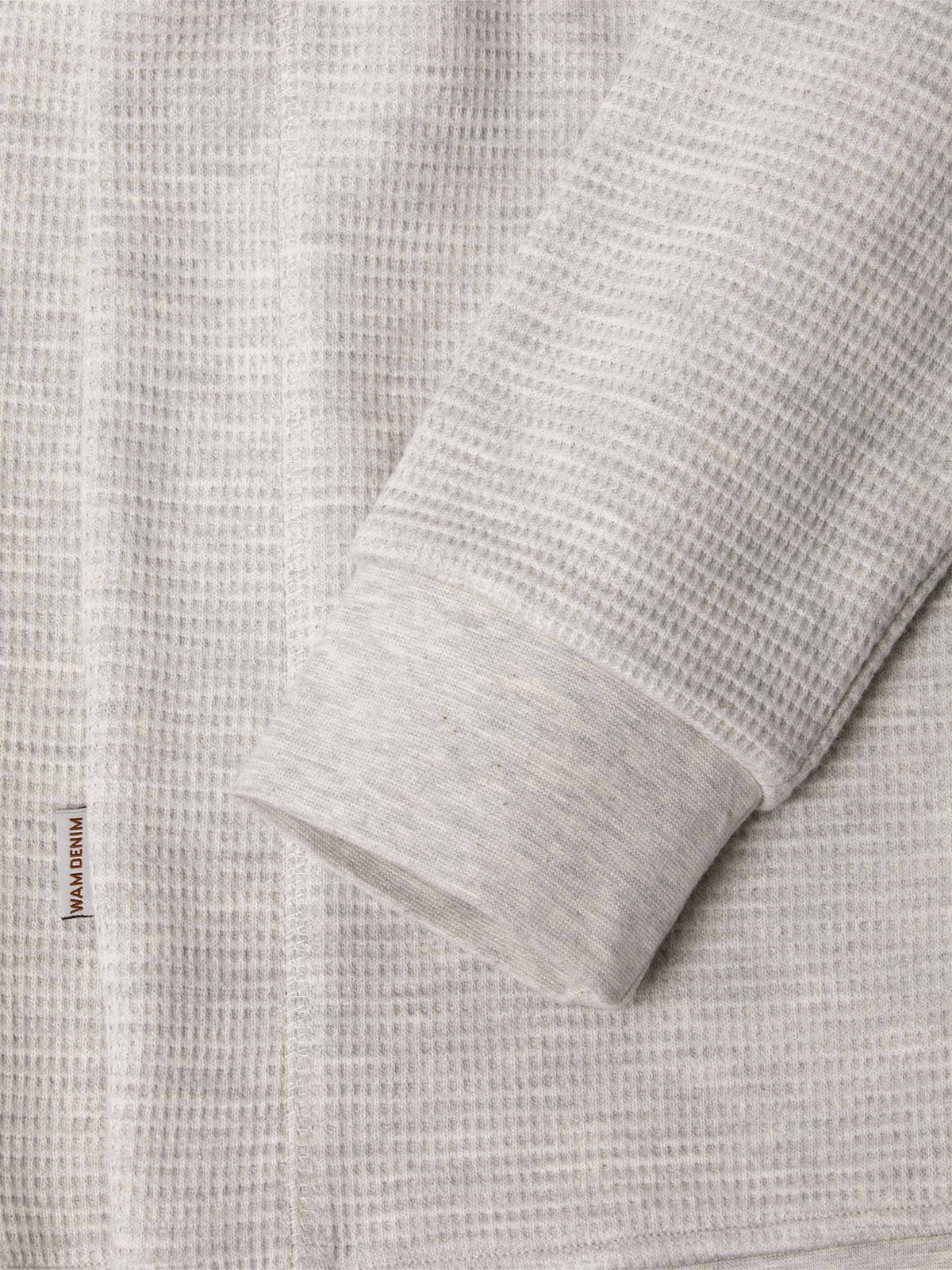 Close-up of light grey waffle-textured sweatshirt sleeve and hem with logo tag – Wam Denim.